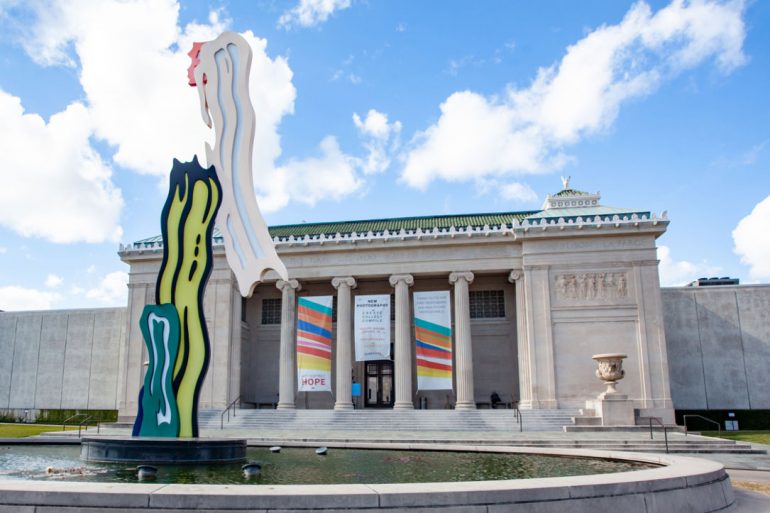 The Neoclassical façade of the New Orleans Museum of Art is visible behind an abstract sculpture by Roy Lichtenstein